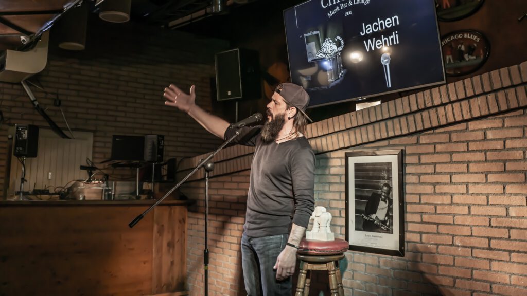 Bearded performer raising one hand while speaking at the microphone