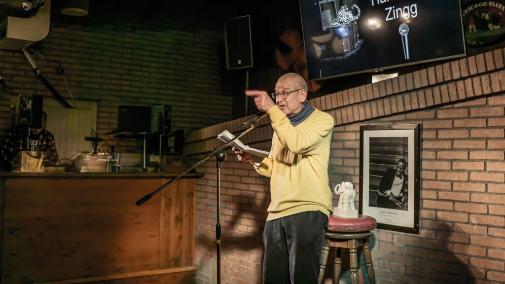 Bald performer in a yellow sweater gesturing while holding a booklet