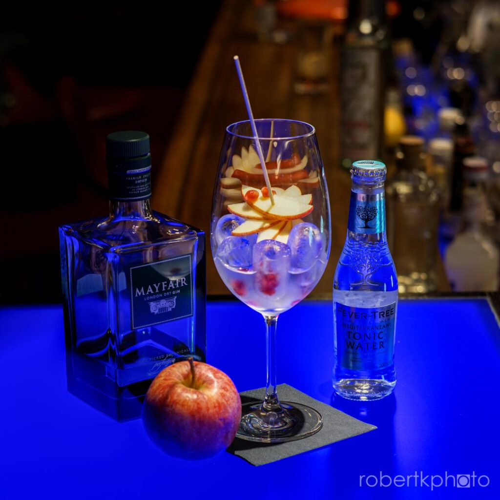 Fruit-garnished gin cocktail with gin bottles on a blue-lit bar