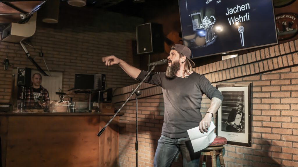 Bearded performer pointing while holding papers at the microphone