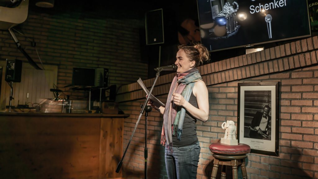 Woman wearing a scarf reading from papers at the Poetry Slam microphone