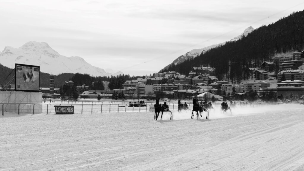 Wide view of skijoring horses crossing the snowy track with St. Moritz and mountains behind