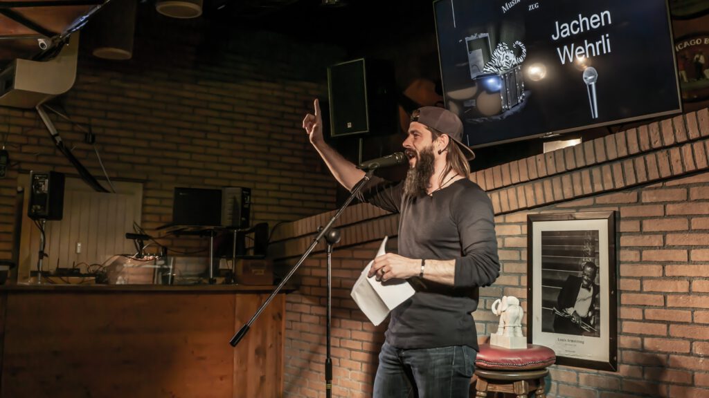 Bearded performer in a cap gesturing while holding papers on stage