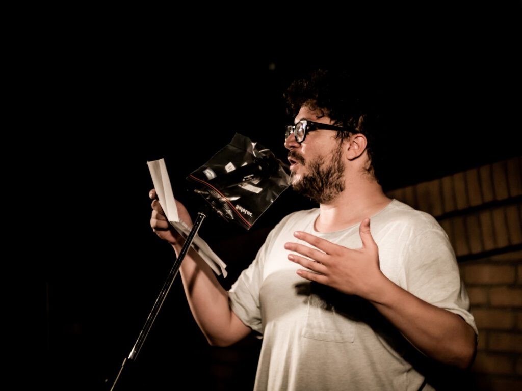 Bearded performer in glasses reading from a booklet at the microphone