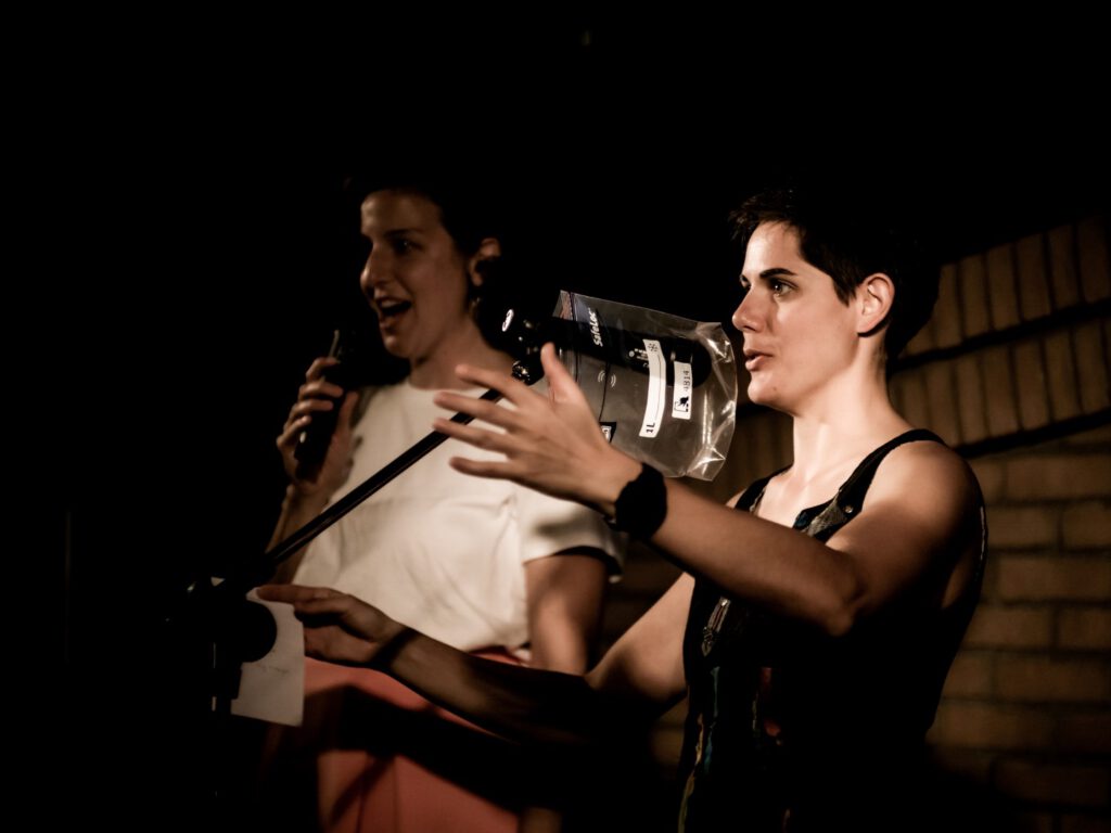 Woman gesturing while speaking beside another performer at Poetry Slam